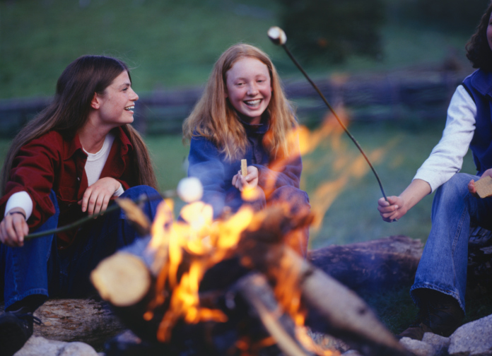 Three teenage girls (16-17) in country field roasting marshmallows over campfire